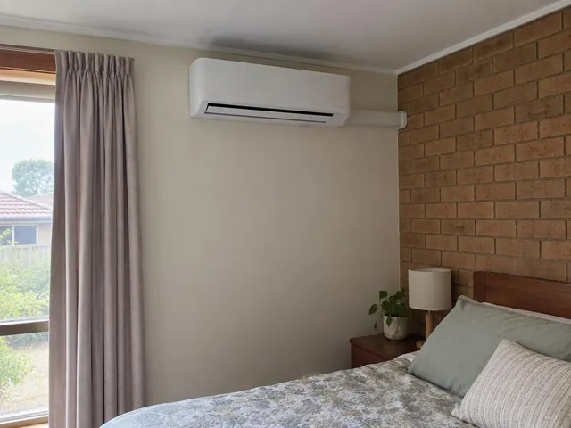 Wall-mounted split system above a bed in a bedroom with brick feature wall and curtains in Elizabeth Grove